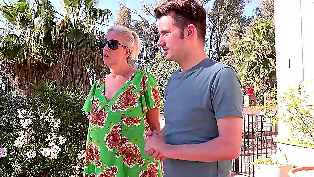 A mature woman with glasses, Maggie Moon, has fun with a young man outdoors.